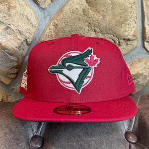 May include: A red baseball cap featuring the Toronto Blue Jays logo. The logo is a stylized blue jay with a green outline and a red maple leaf. The cap has a flat brim and a fitted design.