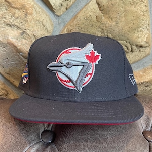 May include: A black baseball cap with a red brim and a team logo featuring a stylized bird and a maple leaf. The cap is set against a textured background.
