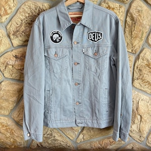 May include: A light blue denim jacket with the text "DEUS" on a patch on the front. The jacket has two chest pockets and two side pockets. A patch with the text "TOO MANY CHIEFS" is on the left chest. The jacket is hanging on a wooden hanger.
