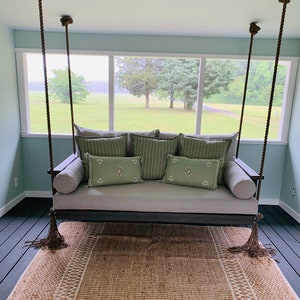 May include: A gray outdoor swing with green and white accent pillows and a matching rug. The swing is suspended by thick brown ropes and tassels. The room has a light blue wall and a view of a green field.