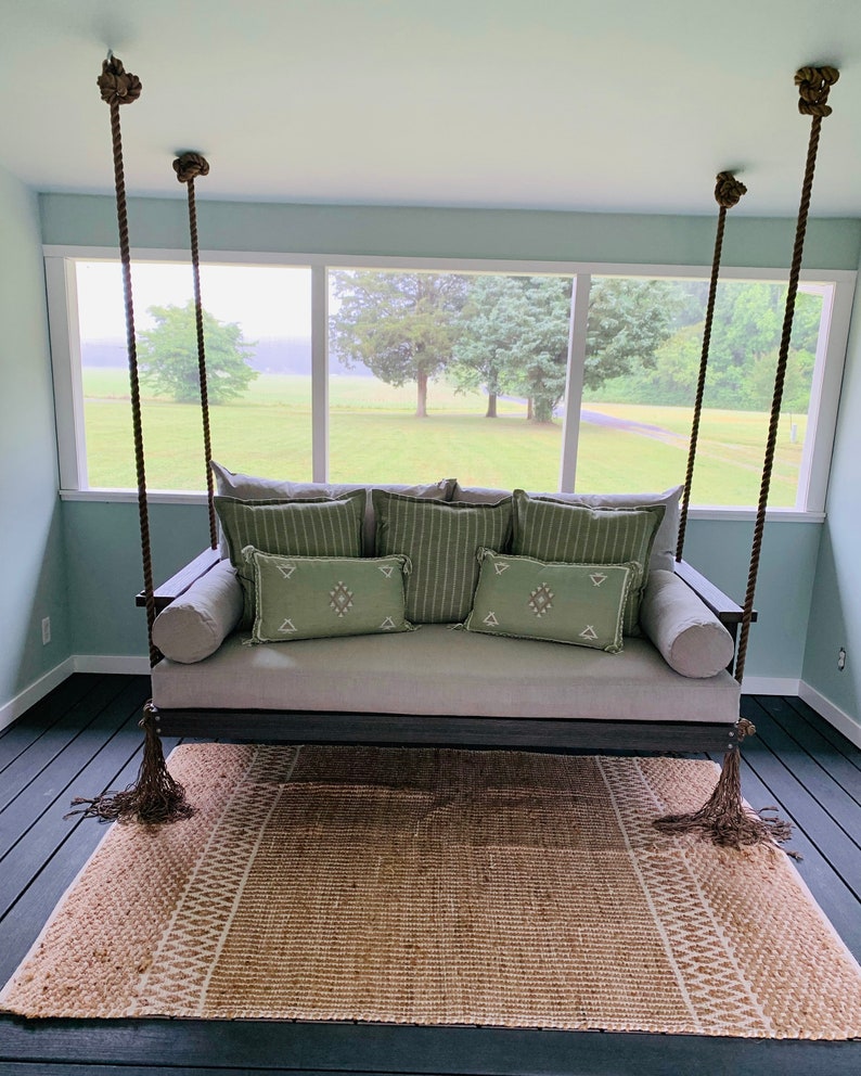 May include: A gray outdoor swing with green and white accent pillows and a matching rug. The swing is suspended by thick brown ropes and tassels. The room has a light blue wall and a view of a green field.