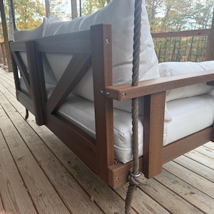 May include: A brown wooden porch swing with white cushions and a rope suspension system. The swing features a slatted back and armrests, and is designed for outdoor use. The swing is suspended from a wooden beam.