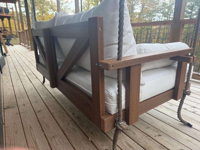May include: A brown wooden porch swing with white cushions and a rope suspension system. The swing features a slatted back and armrests, and is designed for outdoor use. The swing is suspended from a wooden beam.
