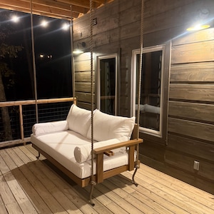 May include: A wooden porch swing with a white cushion and pillows hangs from thick ropes. The swing has a wooden frame and armrests. The porch has wooden decking and a wooden wall in the background.