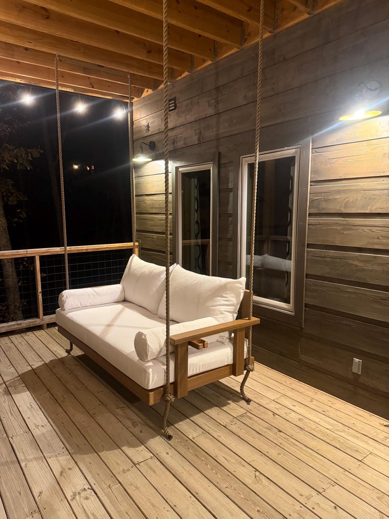 May include: A wooden porch swing with a white cushion and pillows hangs from thick ropes. The swing has a wooden frame and armrests. The porch has wooden decking and a wooden wall in the background.