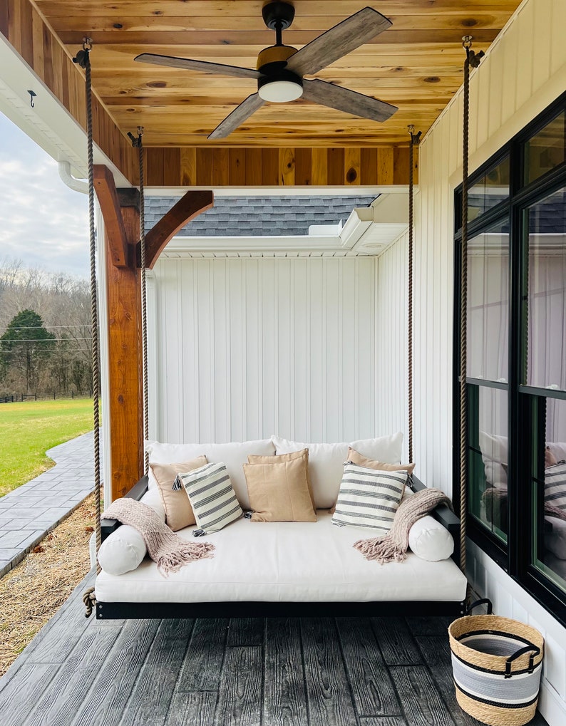 May include: A porch swing with a white cushion and several throw pillows. The swing is suspended by thick ropes from a wooden beam. A ceiling fan is mounted above the swing. The porch has a wooden floor and a woven basket.