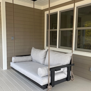 May include: A gray cushioned porch swing with black armrests and a black frame, suspended by thick rope. The swing includes two large back cushions and two bolster pillows. The swing is on a porch with a neutral-colored exterior.
