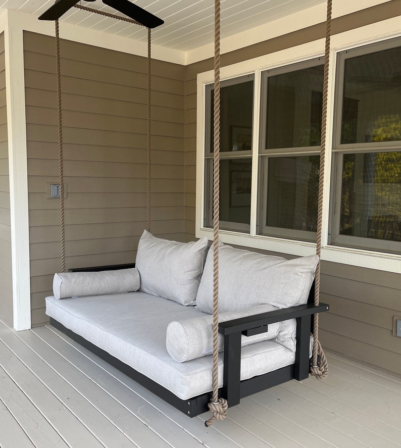 May include: A gray cushioned porch swing with black armrests and a black frame, suspended by thick rope. The swing includes two large back cushions and two bolster pillows. The swing is on a porch with a neutral-colored exterior.