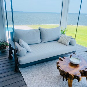 May include: A gray outdoor swing daybed with large pillows and bolster pillows. A wooden side table with a live edge and a decorative bowl are in front of the daybed. The swing hangs from thick rope.