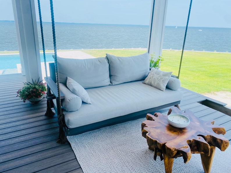 May include: A gray outdoor swing daybed with large pillows and bolster pillows. A wooden side table with a live edge and a decorative bowl are in front of the daybed. The swing hangs from thick rope.