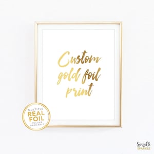 May include: A framed print with gold foil lettering that reads "Custom gold foil print" on a white background. A circular sticker in the lower left corner says "Multiple Real Foil Colors Available". The frame is gold.