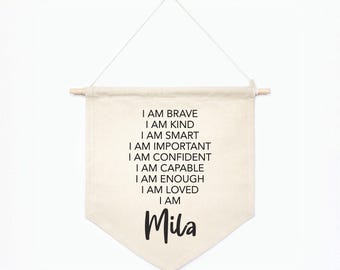 Puede incluir: Un estandarte de pared de tela color crema que cuelga de un palo de madera con un cordel blanco. El texto dice: I AM BRAVE, I AM KIND, I AM SMART, I AM IMPORTANT, I AM CONFIDENT, I AM CAPABLE, I AM ENOUGH, I AM LOVED, I AM Mila, en negro.