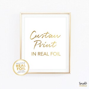 May include: A gold-framed print featuring the gold-coloured text Custom Print IN REAL FOIL. A circular gold badge in the bottom left corner displays the text MULTIPLE REAL FOIL COLORS AVAILABLE. The background of the print is white.