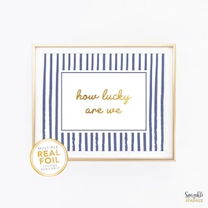 May include: Framed wall art with a gold border. The artwork features a white background with navy blue vertical stripes and the gold text "how lucky are we". A gold sticker in the lower left corner says "Multiple Real Foil Colors Available".