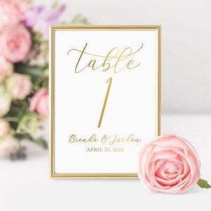May include: A gold framed table number sign with the word "table" and the number "1" in gold script. The names "Brenda & Jordan" and the date "April 25, 2026" are also in gold script. A pink rose is in the foreground.