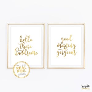 May include: Two framed wall art prints with white backgrounds and gold foil lettering. The left print reads "hello there handsome" and the right print reads "good morning gorgeous" in a cursive script. A gold circular badge reads "MULTIPLE REAL FOIL COLORS AVAILABLE".