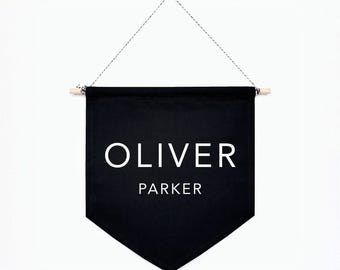 May include: A black fabric wall banner with a pointed bottom edge, featuring the white text OLIVER PARKER printed in the center. The banner is suspended from a light-colored wooden dowel by a thin, black and white twisted cord for hanging.