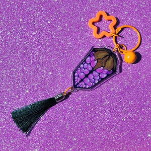 May include: A purple grape keychain with a black tassel and an orange star charm. The keychain is made of clear plastic and has a metal ring.