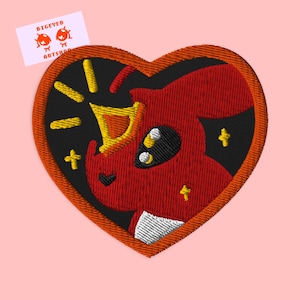 Może przedstawiać: Czerwona, haftowana łatka w kształcie serca z postacią z kreskówki, która nosi koronę i jest otoczona gwiazdami. Postać ma duże oczy i czarny nos.