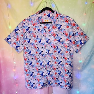 Puede incluir: Camisa de manga corta con un vibrante estampado integral. El diseño presenta pájaros azules, detalles en rojo y elementos florales blancos sobre un fondo azul claro. La camisa está colgada de una percha negra.