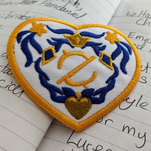 May include: A heart-shaped embroidered patch with a white background, a gold crown, a gold heart, and blue and gold floral designs. The patch is surrounded by a yellow border.