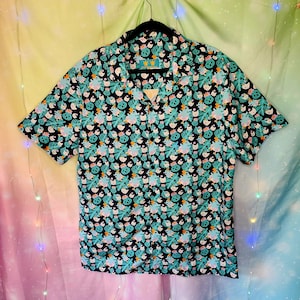 May include: Short-sleeved button-down shirt with a black background and a repeating pattern of teal leaves and cartoon-like characters. The shirt has a collared neckline and is hanging on a black hanger.