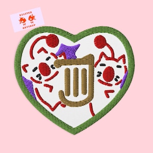 Könnte beinhalten: Ein gestickter Patch in Herzform mit zwei Comicfiguren, einer rot-weißen Harfe und einem lila Stern. Die Figuren haben rote Nasen und tragen rot-weiße Hüte. Der Text "BIGEYED ART SHOP" befindet sich in der oberen linken Ecke.