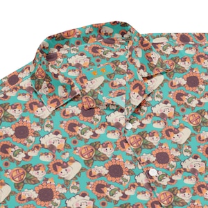 Könnte beinhalten: Ein Hemd mit Knöpfen, das einen türkisfarbenen Hintergrund und ein sich wiederholendes Muster aus Cartoon-Katzen und Sonnenblumen aufweist. Die Katzen sind weiß, orange und braun, und die Sonnenblumen sind gelb und braun. Das Hemd hat einen Kragen und weiße Knöpfe.