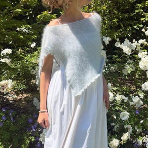 Peut inclure: Un poncho tricoté bleu clair, duveteux, avec un bord festonné, porté sur une robe blanche unie arrivant jusqu'aux pieds. Le poncho présente une maille douce et texturée qui tombe sur les épaules, tandis que la robe a une coupe simple et ample.