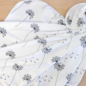 May include: White fabric with a black dandelion pattern. The design features large dandelion seed heads with long stems and individual seeds blowing away. The fabric is draped, suggesting it could be a blanket, scarf, or other textile.