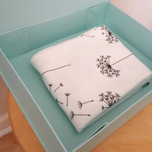 May include: A folded white blanket with a black dandelion design rests inside a light blue box. The dandelion pattern includes seed heads and individual seeds blowing in the wind. The box is open, revealing the blanket.