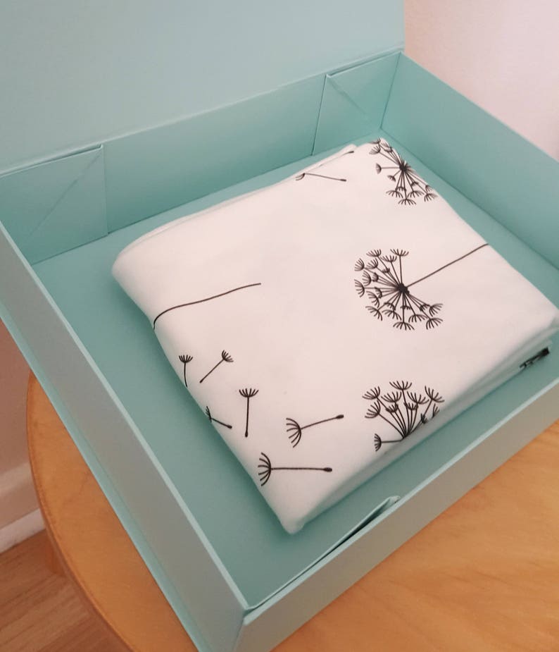May include: A folded white blanket with a black dandelion design rests inside a light blue box. The dandelion pattern includes seed heads and individual seeds blowing in the wind. The box is open, revealing the blanket.