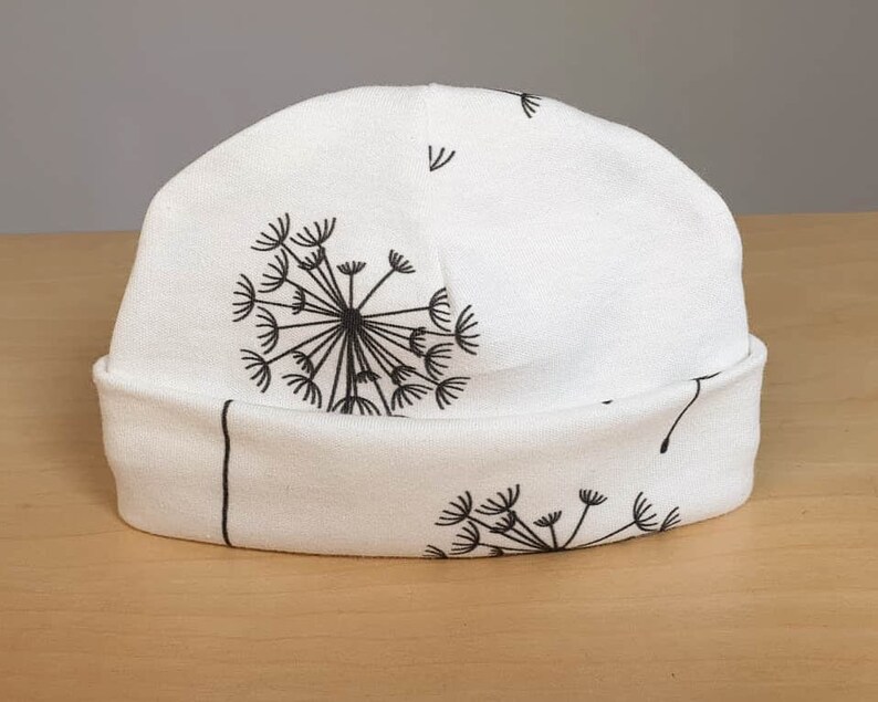 May include: White beanie hat with a black dandelion design. The hat features a rolled-up brim and a repeating pattern of dandelion seed heads. The hat is made of a soft, lightweight fabric.