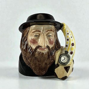 May include: A ceramic mug in the shape of a man's head, wearing a black hat and coat. The handle is decorated with yellow and red dots, and a small pocket watch is attached to the mug.