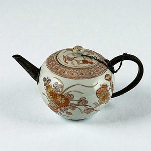 Könnte beinhalten: Kleine, runde Teekanne mit weißem Grund, verziert mit orangefarbenen und braunen Blumenmustern. Deckel und Rand weisen ein ähnliches Design auf. Sie hat einen schwarzen Ausguss und Henkel, die durch eine Kette verbunden sind. Ein kleiner Knopf sitzt auf dem Deckel.