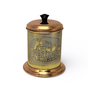 Könnte beinhalten: Ein zylindrischer Behälter mit Deckel und Boden aus Messing und Kupfer. Er hat einen schwarzen Knopf und ein eingraviertes Design an der Seite. Der Behälter ist wahrscheinlich vintage und wird zur Aufbewahrung verwendet. Die Maße sind in Zentimetern.