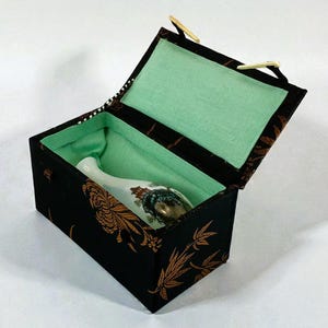 Puede incluir: Una caja negra con un estampado floral dorado y un interior verde claro contiene una botella decorativa blanca y dorada. La caja tiene una tapa con bisagras y un asa pequeña.