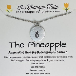 May include: A silver necklace with a pineapple charm. The card reads "The Pineapple, A symbol of Hope for those trying to conceive. Like the pineapple, your tough outer shell protects your sweet core from life's struggles. But being tough is hard. Just remember: You are brave. You are strong. You are enough. You are never, ever alone."