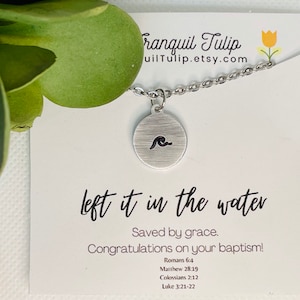 May include: A silver necklace with a wave charm and a card that says "left it in the water. Saved by grace. Congratulations on your baptism!" with bible verses listed below.