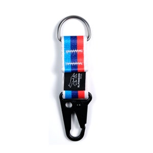 Pu&ograve; includere: Portachiavi bianco con un moschettone nero e un cinturino a strisce blu, rosso e bianco. Il cinturino ha la scritta "ALPINIHOUND PET CO." stampata su di esso.