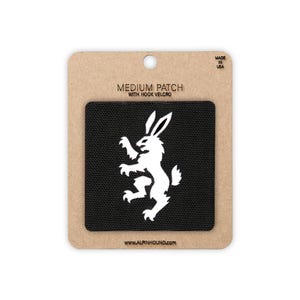 Peut inclure: Un écusson carré noir avec un motif blanc représentant un lapin debout. L'écusson est fixé sur un support en carton brun rectangulaire portant l'inscription MEDIUM PATCH WITH HOOK VELCRO, WWW.ALPINHOUND.COM et MADE IN USA en haut à droite.