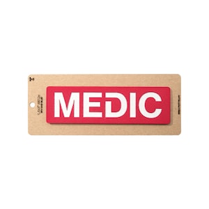 May include: A red rectangular patch with the word "MEDIC" in bold white letters. The patch is 1.5 x 5 inches and has hook velcro. The patch is attached to a brown cardboard backing.