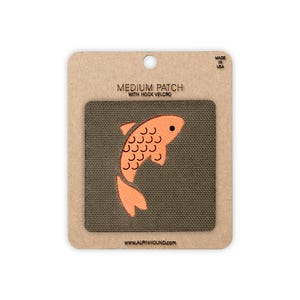May include: A square olive green patch featuring an orange fish graphic with black scale outlines. The patch is mounted on a rectangular cardboard backing labeled MEDIUM PATCH WITH HOOK VELCRO, MADE IN USA, and WWW.ALPINHOUND.COM.