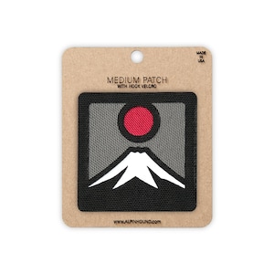 May include: A square patch with hook velcro backing on a cardboard display card. The patch features a gray background, a white mountain silhouette, and a red circular sun. Text on the card reads MEDIUM PATCH WITH HOOK VELCRO, MADE IN USA, and www.ALPINHOUND.com.