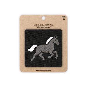 May include: A square black patch featuring a grey horse with a white mane and tail. The patch is mounted on a rectangular tan cardboard backing labelled MEDIUM PATCH WITH HOOK VELCRO, MADE IN USA, and the website address WWW.ALPINHOUND.COM.