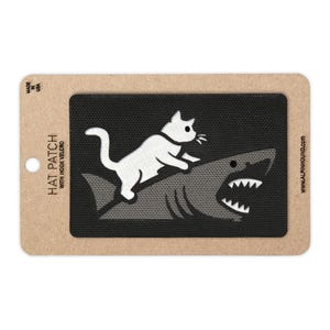 May include: A black rectangular hat patch with a white cat riding a gray shark. The patch is on a cardboard backing with the text "HAT PATCH WITH HOOK VELCRO" and "MADE IN USA". The website www.ALPINHOUND.com is also visible.
