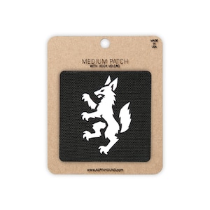 May include: A black square patch featuring a white graphic of a standing wolf. The patch is mounted on a brown cardboard backing labeled MEDIUM PATCH WITH HOOK VELCRO, MADE IN USA, and WWW.ALPINHOUND.COM.