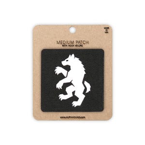 May include: A square black patch featuring a white silhouette of a wolf standing on its hind legs. The patch is mounted on a tan cardboard backing labelled MEDIUM PATCH WITH HOOK VELCRO and WWW.ALPINHOUND.COM, with MADE IN USA printed at the top.
