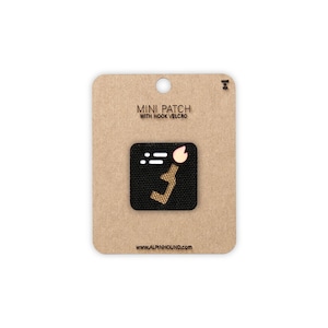May include: A mini patch with hook velcro attached to a cardboard backing. The black patch features a stylized design with a light brown shape and a pink accent. The text "MINI PATCH WITH HOOK VELCRO" is printed above the patch.