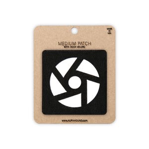 May include: A black medium patch with hook and loop fastening, featuring a white geometric design. The patch is on a cardboard backing with the text "MEDIUM PATCH WITH HOOK VELCRO" and "MADE IN USA". The website address www.alpinhound.com is also visible.